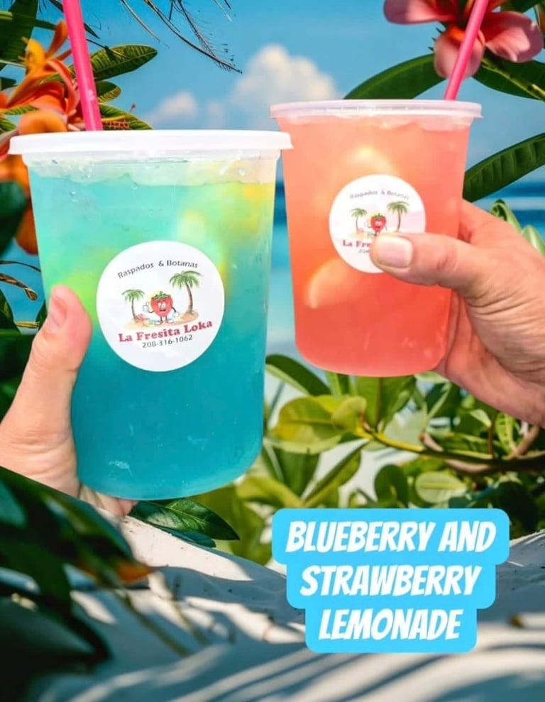 Two tropical drinks held outdoors: blue layered blueberry lemonade and pink strawberry lemonade from La Fresita Loka with pink straws and flower labels